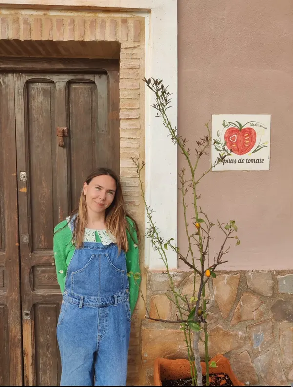 Irene, con peto vaquero y jersey verde, sonríe junto a la puerta de madera de Pepitas de tomate, al lado del cartel y de un limonero en maceta
